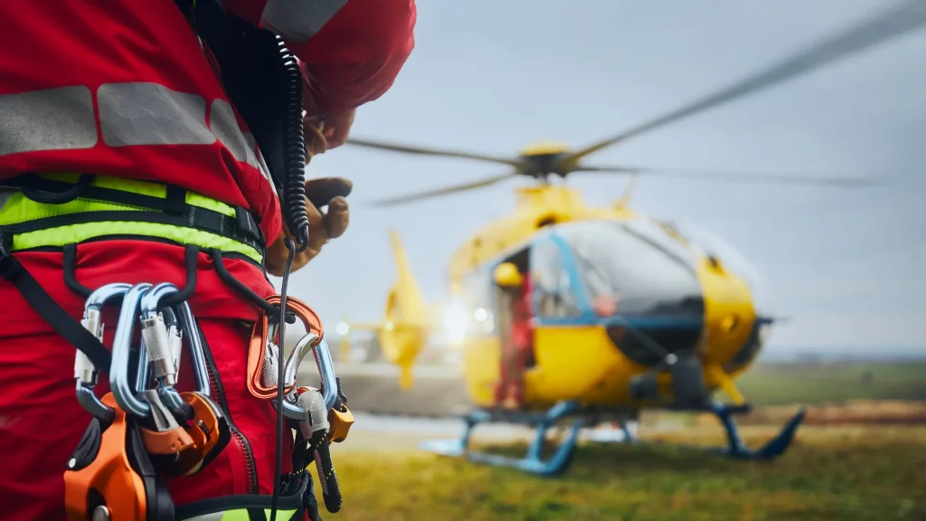 A close up photo of a paramedic in red with carabeaners at their hip as a yellow paramedic helicopter lands in an emergency response.