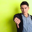 A man holding their thumb down in front of a neon yellow green color. He wears a blue button-up shirt with a cardigan over it.