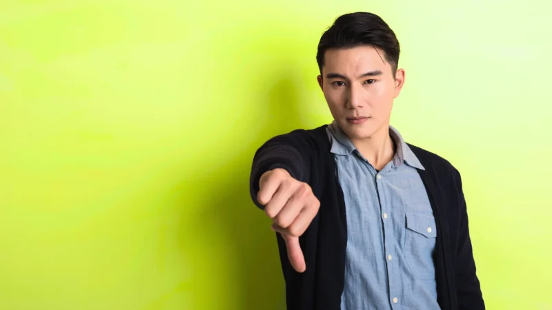 A man holding their thumb down in front of a neon yellow green color. He wears a blue button-up shirt with a cardigan over it.