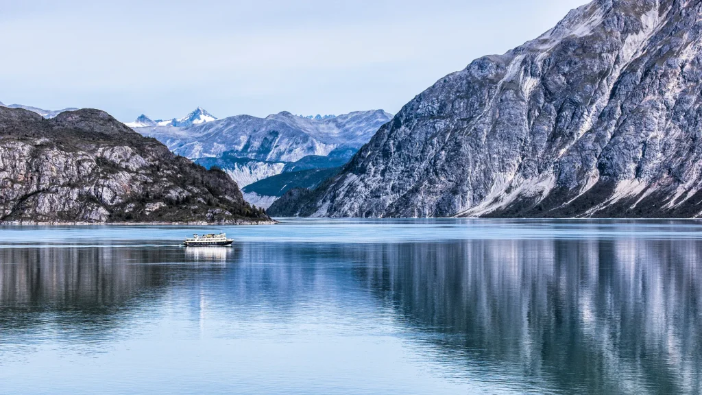 A ferry boat along the Alaska Marine Hightway. Large rocky and snowy landscapes surround the ferry and the glass like water it floats atop.