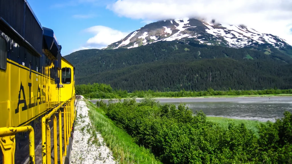 A black and yellow Alaskan train with "Alaska" on the side chugging along a body of water as a clouded mountain juts out behind it.