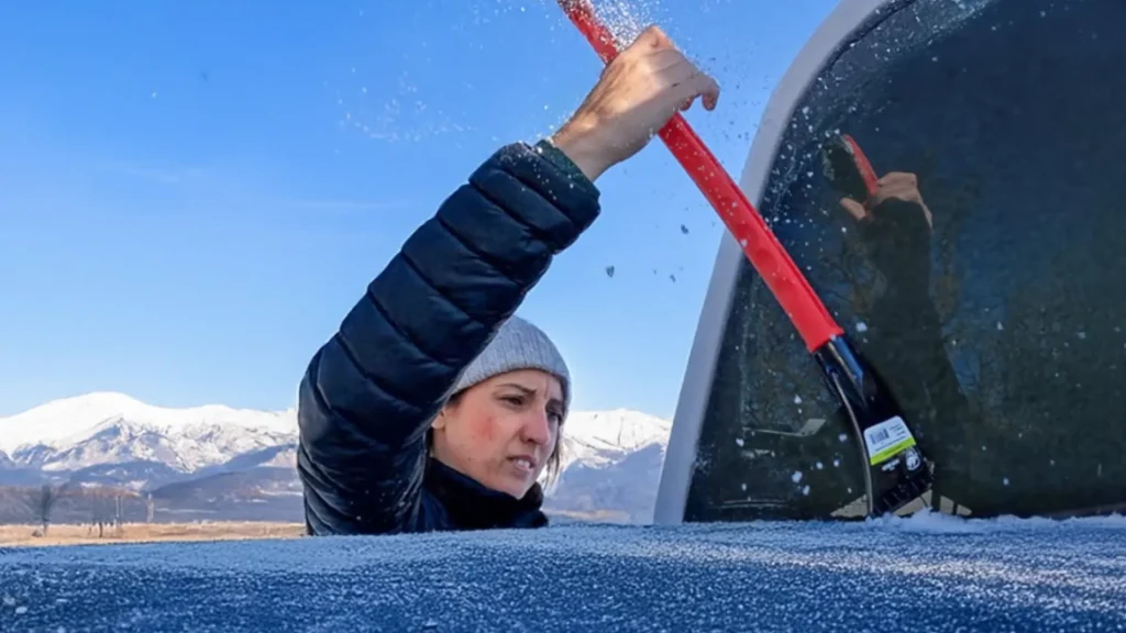HappilyEverHanks using an ice scraper to remove ice off of the rear windshield of their truck, as mountains take up the background along with a bright blue sky. 