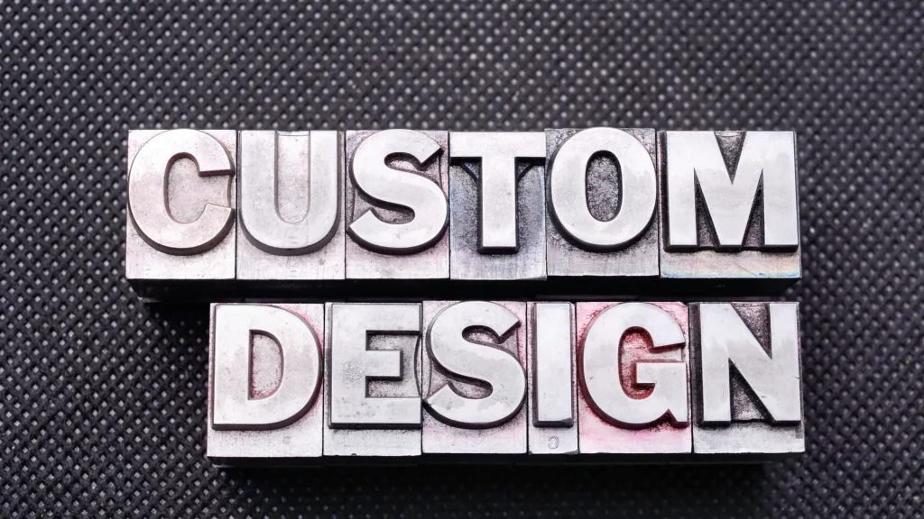 In metal embossing tools, "CUSTOM DESIGN" is spelled out against a black rubber mat background.