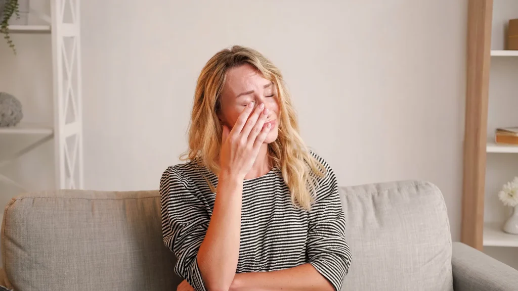 A woman emotionally upset as she holds her hand to her face while seated on a sofa. Behind her is a white wall with some wooden shelving. She wears a black and white striped long sleeve shirt.
