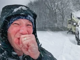 HappilyEverHanks holding his hand up, while taking a selfie as it continues to snow around him while showing the RV and Truck covered in snow.