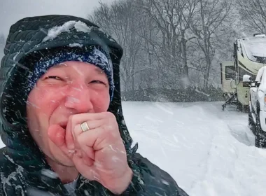 HappilyEverHanks holding his hand up, while taking a selfie as it continues to snow around him while showing the RV and Truck covered in snow.