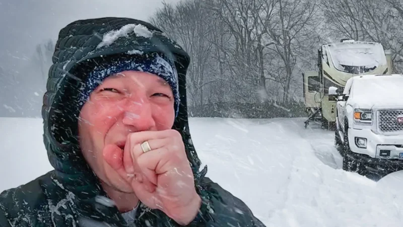 HappilyEverHanks holding his hand up, while taking a selfie as it continues to snow around him while showing the RV and Truck covered in snow.