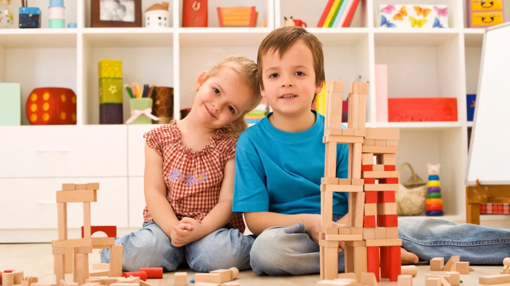 Two kids sitting beside each other in a playroom smiling as an array of wooden blocks lay before them on the floor and in stacks forming towers.