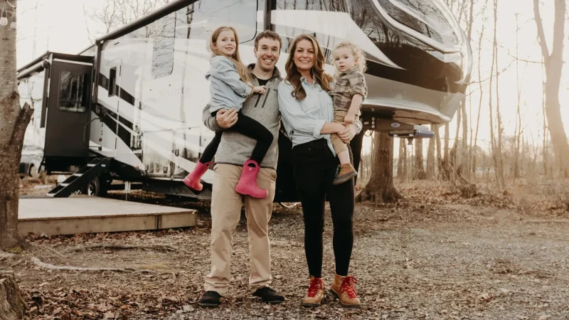 Nathan and Marissa from Less Junk, More Journey with their two children as they stand holding them on their hips in front of their RV.