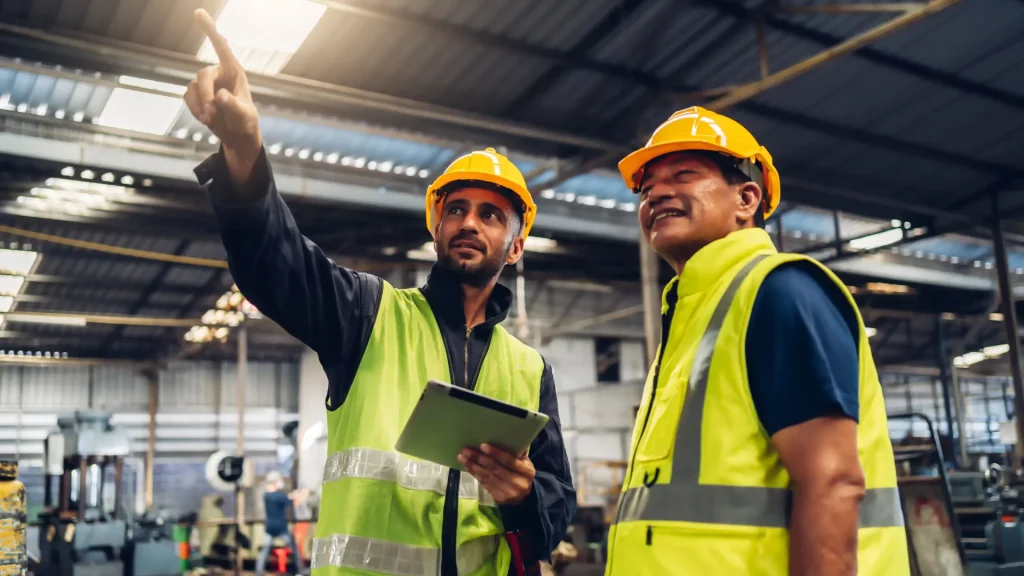 Two manufacture workers in a warehouse wearing bright yellow vests and golden yellow safety helmets as one points off in the distance and the other looks.