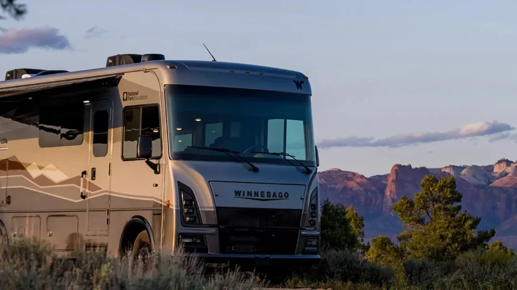 Winnebago motorhome in front of a rocky ridge with a dusky grey blue sky and thin wispy clouds in the background.