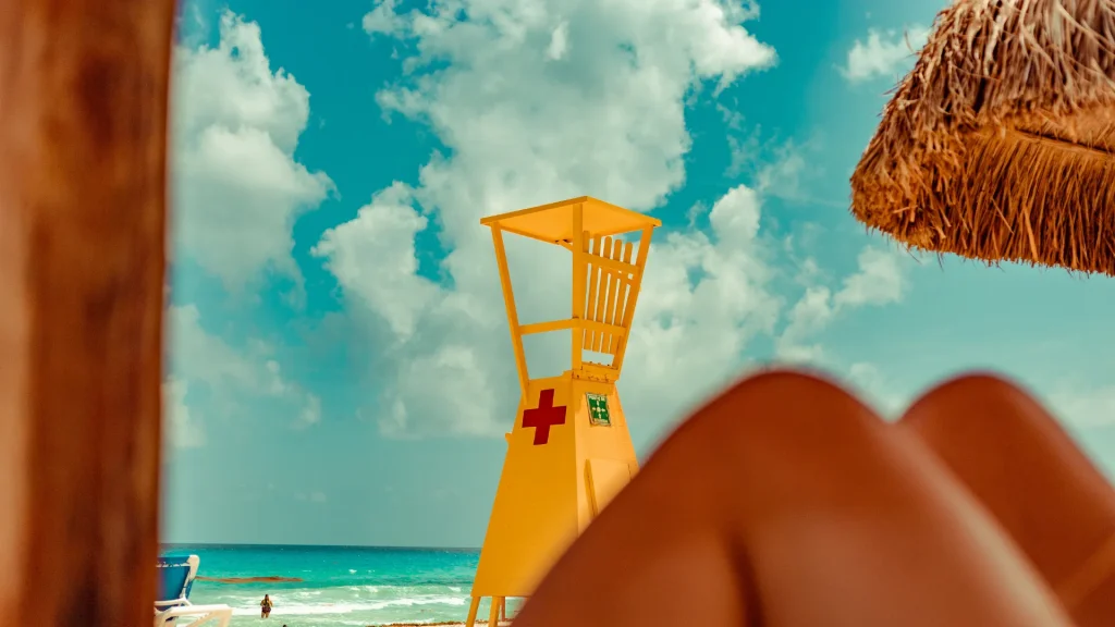 A pair of knees sits blurry in the foreground as the camera focuses on a yellow lifeguard station on the beach. The bright blue sky is spotted with large puffy clouds and water is a bright turquoise blue. 