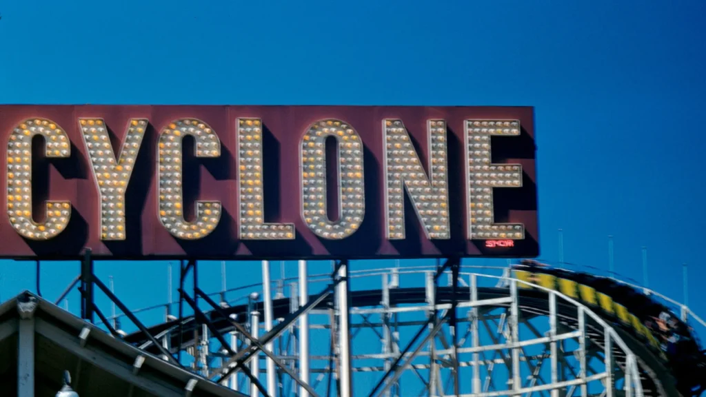 The Cyclone's sign from the Revere Amusement park before it was demolished. The sign is partially blurry as it sits against a bright blue clear sky while one of the coaster cars speeds by.