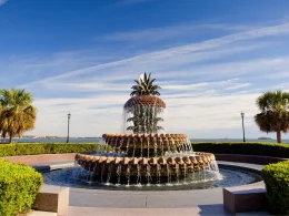 The infamous Pineapple Fountain in Charleston SC symmetrically displayed with shrubs and walkways around and beside it with a blue sky containing wispy clouds.