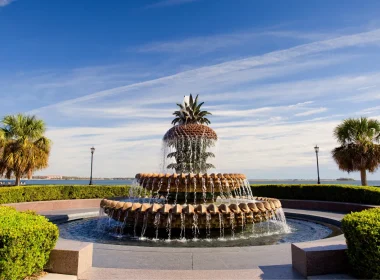 The infamous Pineapple Fountain in Charleston SC symmetrically displayed with shrubs and walkways around and beside it with a blue sky containing wispy clouds.