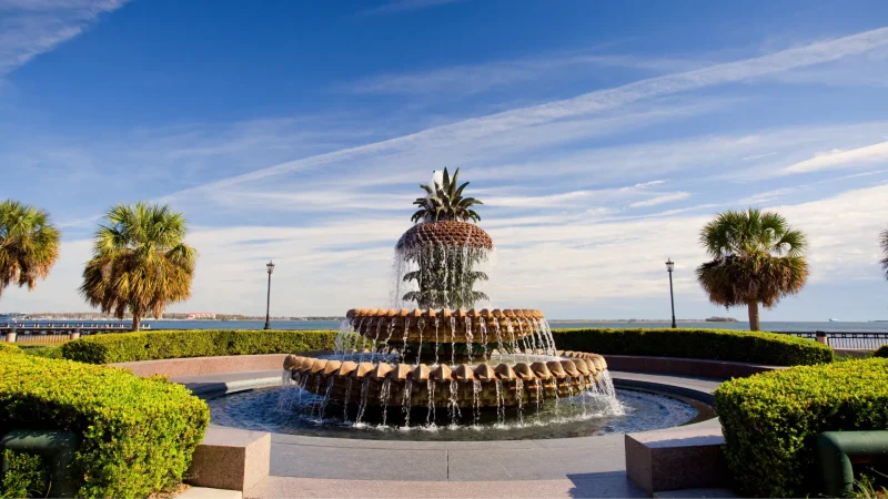 The infamous Pineapple Fountain in Charleston SC symmetrically displayed with shrubs and walkways around and beside it with a blue sky containing wispy clouds.