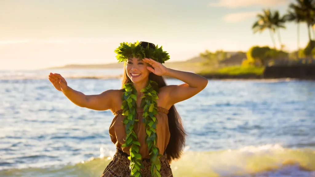 A Hawaiian Hula dancer dancing on the beach of Kauai Hawaii at sunset. The sun casts a golden glow on her and the land. Water and a stretch of the island sit behind her as she dances. 