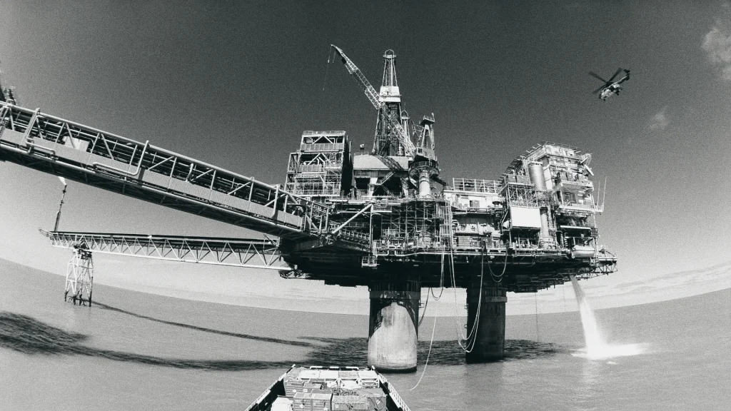 A black and grey photo of an old off-shore oil rig. A helicopter flies over in the clear sky.