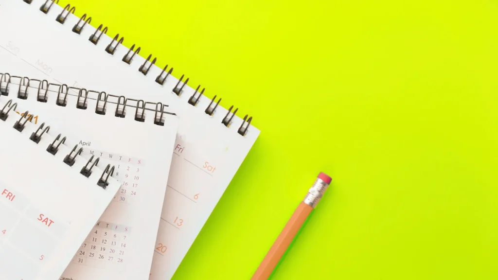 A neon yellow-green background, three notepads of calendars or schedules lay atop each other as a traditional golden pencils lays beside them. 