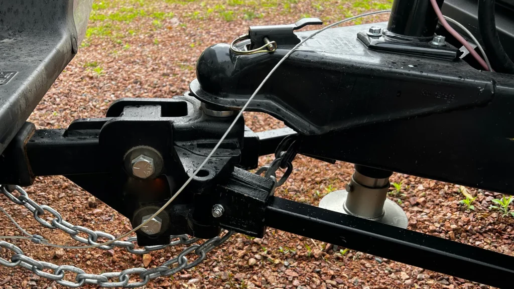 An RV Trailer Hitch hooked up to a ball hitch on a truck.  The materials are all black and silver as we see the gravel beneath the truck and trailer. 
