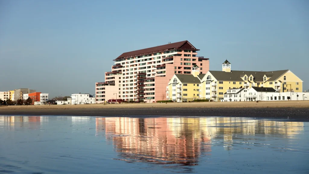Revere Beach is a public beach in Revere, Massachusetts. It was founded in 1895 as the first public beach in the U.S. Pastel buildings sit on the beach with a blue-grey sky in the backdrop. The buildings are reflected in the water.