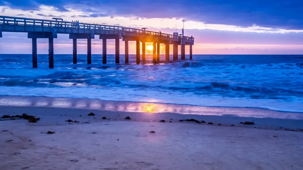 St. Augustine Pier on St. Augustine Beach in Florida at sunset. The sun is dipping below the horizon as everything turns dark blue and purple at dusk while the sun eminates an orange light between legs of the pier.