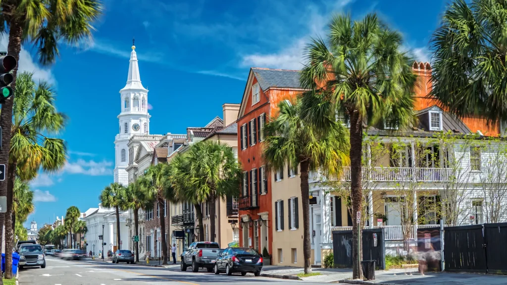 The Charleston Historic District displayed in a street photo of St. Michaels Church and Broad St. in Charleston, SC. Palms line up and down the road as cars sit parked ont he side of the street. 