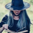 A woman in a witche's hat outside, holding a book as she pages through it.