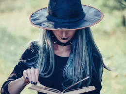 A woman in a witche's hat outside, holding a book as she pages through it.