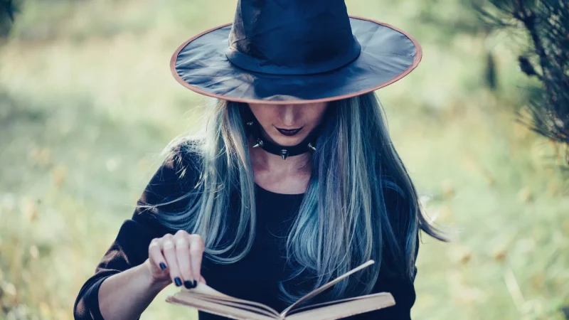 A woman in a witche's hat outside, holding a book as she pages through it.