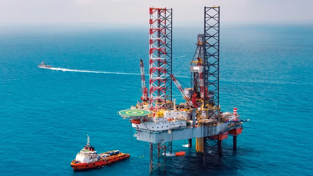 An aerial of an offshore drilling site with a helicopter pad and two boats in the water. One speeds off behind the drilling site as the other sits stationary next to it. 
