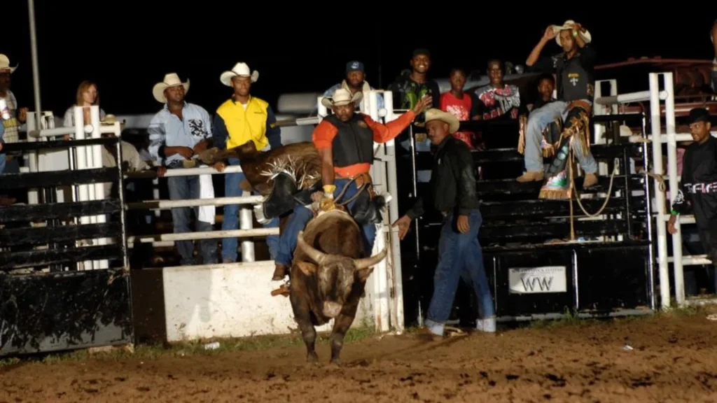 An African American man on a bull riding with their hand in the air as onlookers watch.