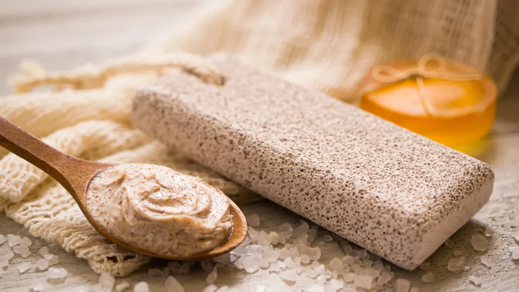 A skin scrub product is portioned onto a wooden spoon set atop a terry cloth and rock salt beside a pumice stone and a round orange bar of soap tied with a hemp ribbon. 