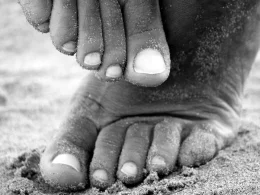 An up close black and white photo of feet in the sand on a beach as one rubs the other.