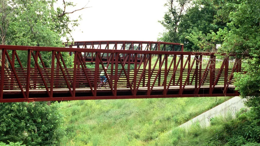 A photo from within Gwynns Falls Trail of a red bridge that has a cyclist riding across it.