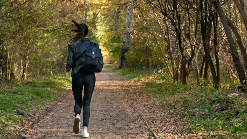 A person running down a trail in a densely forested area with leaves on the the ground. 