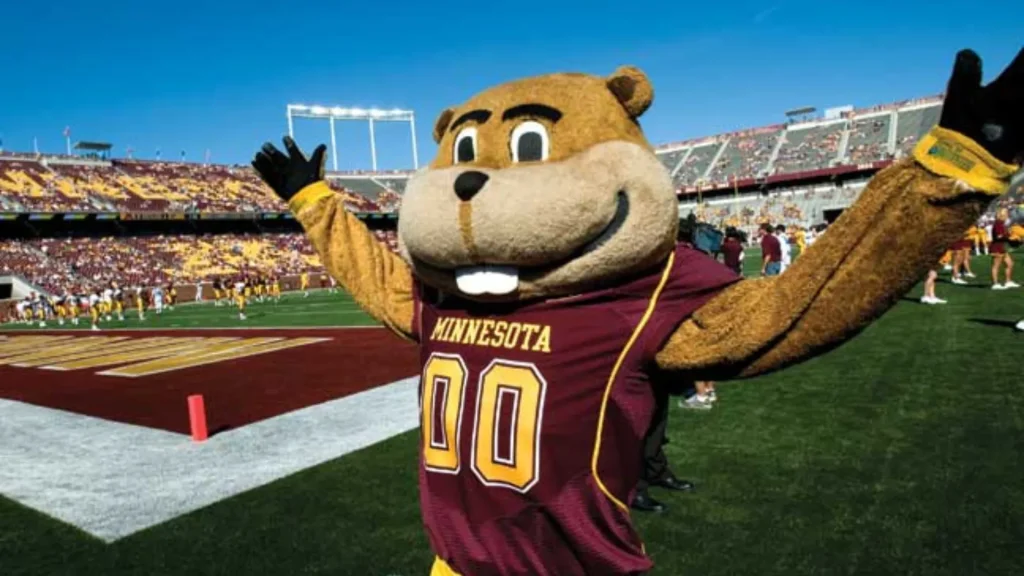 The University of Minnesota's mascot Goldy the Gopher in the stadium surrounded by football players and fans alike.