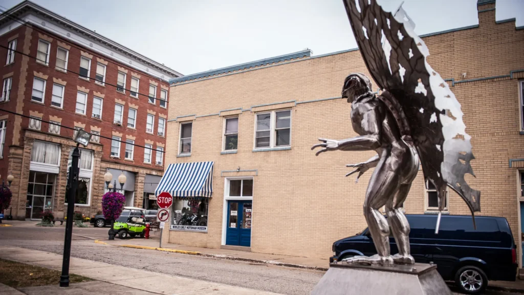 The mothman statue, brightly chrome in color as it stand beside a sidewalk in a town.