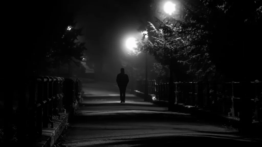 A black and grey photo of a hooded figure walking at night through a foggy walking path.