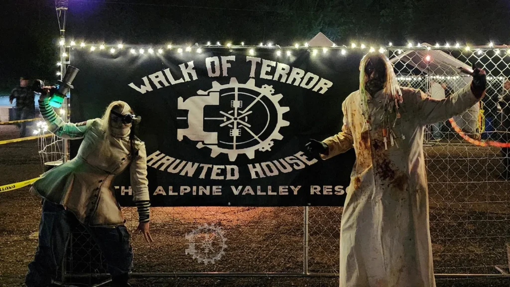 A black canvas sign with "Walk of Terror Haunted House The Alpine Valley Resort" in white text on it. Two people dressed up in scary costumes stand on each side of it as it is hung on a chain link fence. 
