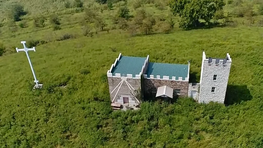 An aerial photo of the Metamora Castle in an open field. It has a simple structure with a large white cross standing beside it.
