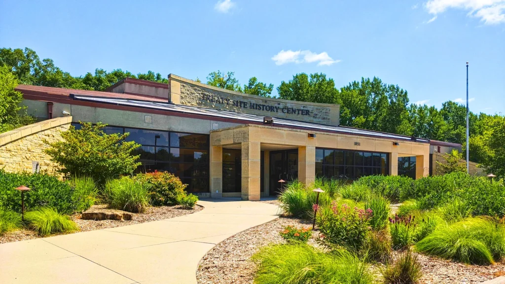 A photograph of the outside of the Nicollet County Historical Society Museum with neatly manicured landscaping in front of it. It's a sunny bright blue sky day with few clouds in the sky.