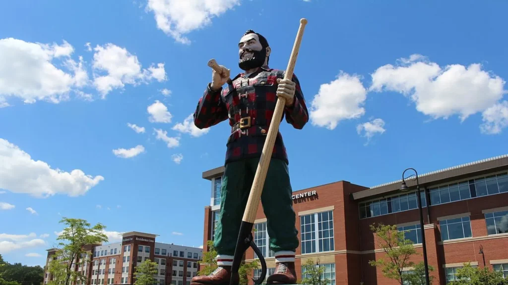 The Paul Bunyan Statue in Bangor, Maine. The photo depicts a sunny day with a bright blue sky dotted with clouds. as city buildings stand behind the statue.