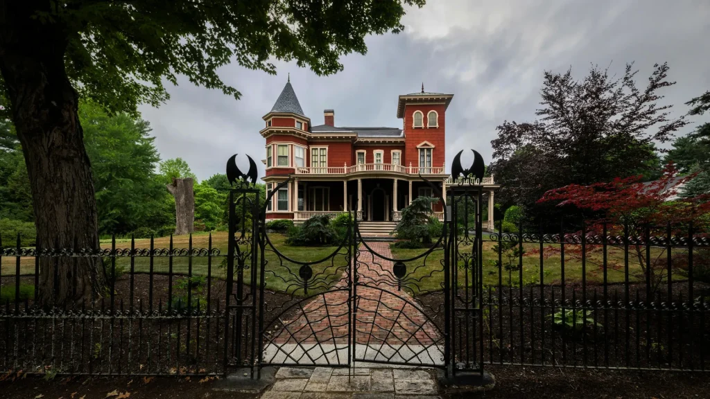 The Stephen King House is a Victorian-style home with a red brick exterior. There is a black iron gate with spiders and bats in the ironwork. The lawn is neatly manicured, and the sky is grey.