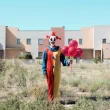 A clown in an arid environment stands in an open field in front of a town with a pale, clear blue sky, holding red balloons.