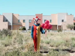 A clown in an arid environment stands in an open field in front of a town with a pale, clear blue sky, holding red balloons.