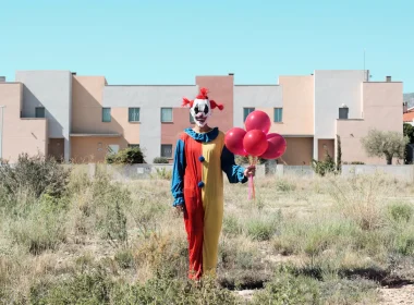 A clown in an arid environment stands in an open field in front of a town with a pale, clear blue sky, holding red balloons.
