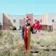 A clown in an arid environment stands in an open field in front of a town with a pale, clear blue sky, holding red balloons.
